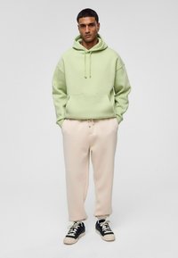 Lichtgroene hoodie met trekkoorden en een voorkeurszak, gecombineerd met beige joggers. Zwarte en crèmekleurige sneakers maken de outfit compleet.