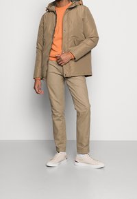 Chaqueta aislante beige con capucha, suéter naranja debajo, chinos de color beige claro y zapatillas de crema. Textura suave, diseño minimalista.