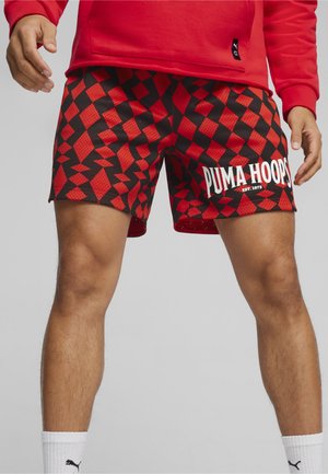 Jambes d'homme portant un short de basketball Puma Hoops à motifs géométriques rouges et noirs et des chaussettes blanches Puma, debout les mains détendues le long du corps.