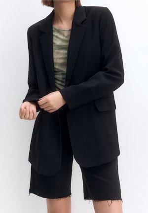 Person trägt einen schwarzen oversized Blazer über einem grünen gemusterten Top und schwarzen Shorts mit ausgefranstem Saum und richtet den Blazerärmel.