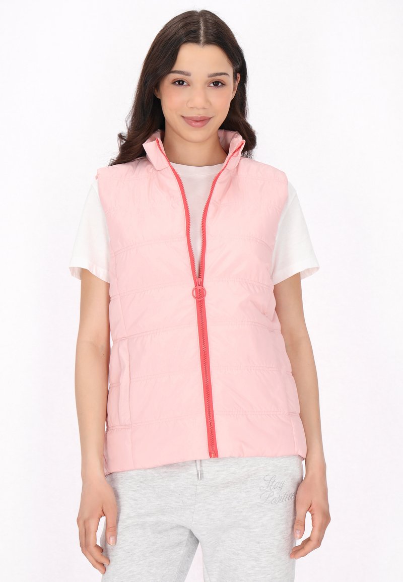 Gilet imbottito leggero rosa con colletto alto e zip frontale. Accentuato da una zip corallo brillante. Indossato sopra una maglietta bianca.