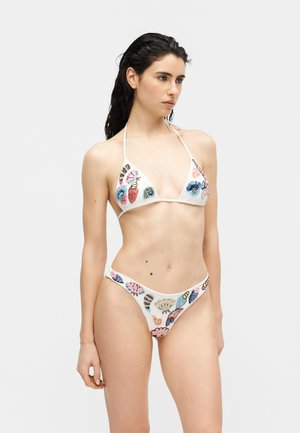 Γυναίκα που φοράει λευκό μπικίνι με πολύχρωμη κεντητική με κοχύλια και θαλάσσια πλάσματα, στέκεται μπροστά σε απλό ανοιχτό γκρι φόντο.