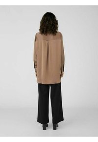 Donna con capelli ricci che indossa una lunga blusa beige e pantaloni neri a gamba larga, in piedi di spalle su uno sfondo semplice.