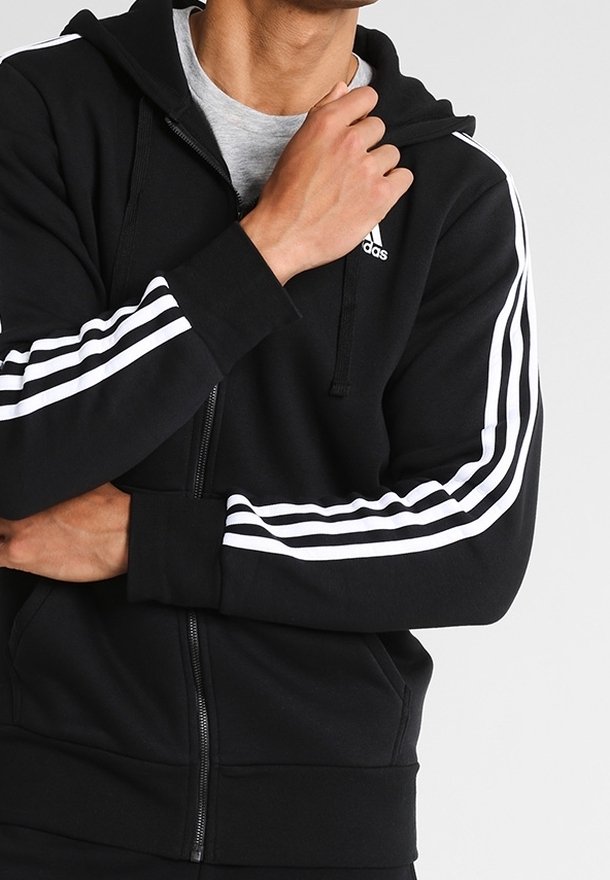 Homme portant un sweat à capuche zippé noir Adidas avec des bandes blanches sur les manches et un t-shirt gris clair en dessous, main près du col.