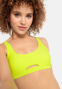 Neon zelený bralet s okrúhlym výstrihom a strihom v prednej časti, vyrobený z hladkého a pružného materiálu.