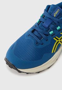 Blå sportssko med teksturert overdel, med grønne og gule detaljer og gummisåle. Designet inkluderer lisser og forsterket tåområde.