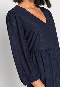 Robe bleu marine en tissu léger et texturé avec un décolleté en V, des manches bouffantes et un ourlet évasé avec des détails en œillets.