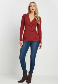 Burgunderrotes Langarmshirt mit V-Ausschnitt und Wickeldesign, kombiniert mit blauen Skinny-Jeans und braunen Stiefeletten. Weicher Stoff mit taillierter Silhouette.