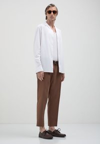 Uomo con occhiali da sole arancioni, camicia bianca sbottonata sopra una canotta bianca, pantaloni corti marroni e scarpe stringate marroni scure in piedi contro uno sfondo neutro.