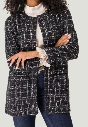 Femme portant une veste ouverte texturée à carreaux noir et blanc sur un chemisier crème avec des poignets à volants, associée à un jean bleu foncé, les bras croisés.