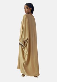 Lang, flydende beige kaftan lavet af glat stof, med brede ærmer og en dekorativ kant ved manchetterne.