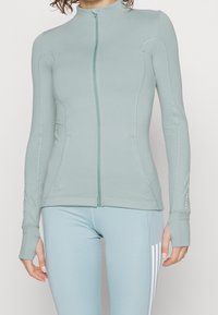 Světlé zelená sportovní bunda na zip, vyrobená z hladké látky, s bočními kapsami, otvory na palce v rukávech a dekorativními švy.