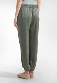 Luźne spodnie dresowe w oliwkowej zieleni, wykonane z lekkiego materiału, z elastycznym pasem i mankietami. Noszone z jasnoróżowymi butami.