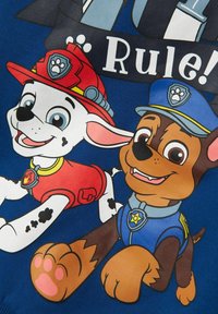 La maglietta blu presenta due cani dei cartoni animati: un cane bianco con un elmetto da pompiere rosso e un cane marrone con un cappello da poliziotto blu, entrambi sorridenti e in corsa.