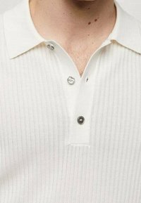 Polo blanc côtelé avec une patte boutonnée ornée de boutons argentés, un col classique et un tissu texturé.
