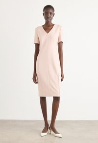 Pink fitted dress with short sleeves, V-neckline, and vertical seams. Smooth texture, knee-length skirt, paired with white pointed-toe heels.