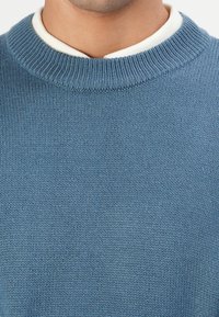 Close-up van een persoon die een lichtblauwe gebreide trui over een witte ronde hals t-shirt draagt, met de nadruk op de nek en het bovenste gedeelte van de borst.