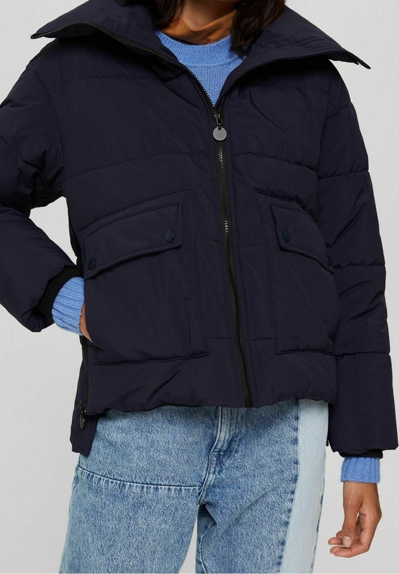 Person iført en sort quilted jakke med store frontlommer, blå strikketrøje og tofarvede blå jeans mod en ensfarvet baggrund.