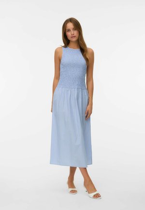 Kvinde iført blå, ærmeløs, smocket topkjole med stribet, flydende nederdel og hvide åbne tå-hæle, stående mod en ensfarvet hvid baggrund.