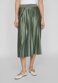 Gonna midi plissè verde in tessuto lucido con una ampia vita elastica. Indossata con scarpe basse color crema con una leggera lucentezza.