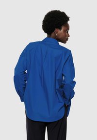 Femme avec des cheveux courts et bouclés portant une chemise ample à manches longues bleue et un pantalon sombre, de profil avec les mains dans les poches.