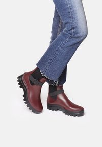 Botas de tobillo impermeables burdeos con suela negra gruesa, correa ajustable y parte superior de goma, combinadas con jeans azules de dobladillo deshilachado.