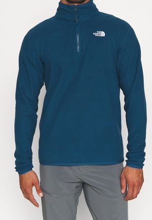 Hombre con un jersey polar azul con cremallera hasta el pecho y el logo de The North Face en el pecho, y pantalones grises, de pie frente a un fondo liso.