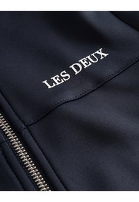 Navy zip-up jakke laget av glatt stoff, med hvit tekst "LES DEUX" og sølv glidelåsbeslag.