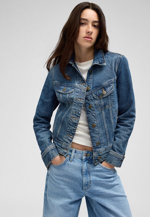 Junge Frau mit langen braunen Haaren, die eine blaue Jeansjacke über einem weißen Top und hellblauen Jeans trägt, Hände in den Taschen, neutraler Hintergrund.
