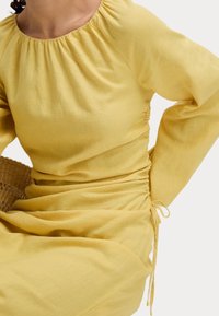 Gelbes Leinenkleid mit langen Ärmeln, gerafftem Rundhalsausschnitt und seitlichem Raffung. Strukturierte Stoffdetails betonen die fließende Silhouette.
