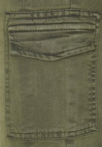 Olive zelená denimová tkanina s jednou zadní kapsou a detaily přesného šití. Textura je hladká s jemným diagonálním vzorem.