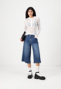 Top alb cu dantelă, cu mâneci lungi și model floral, asortat cu culottes din denim albastru cu picioare largi și pantofi chunky negri cu șosete albe. Geantă neagră.