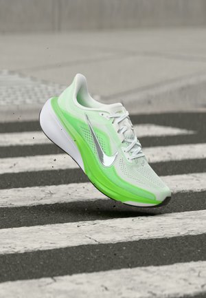 White and neon green Nike running shoe with silver swoosh, mid-air over zebra crossing, with small debris kicked up from ground.