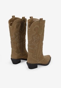 Botas de vaquero de ante marrón con costuras decorativas, punta afilada y tacón bajo. Incluyen lengüetas para facilitar su uso.