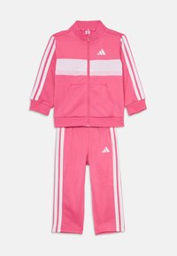 Rožinė Adidas treningo apranga, kurią sudaro zip-up striukė su baltais juostų akcentais ir atitinkamos kelnės, pagamintos iš lygios, elastinės medžiagos.