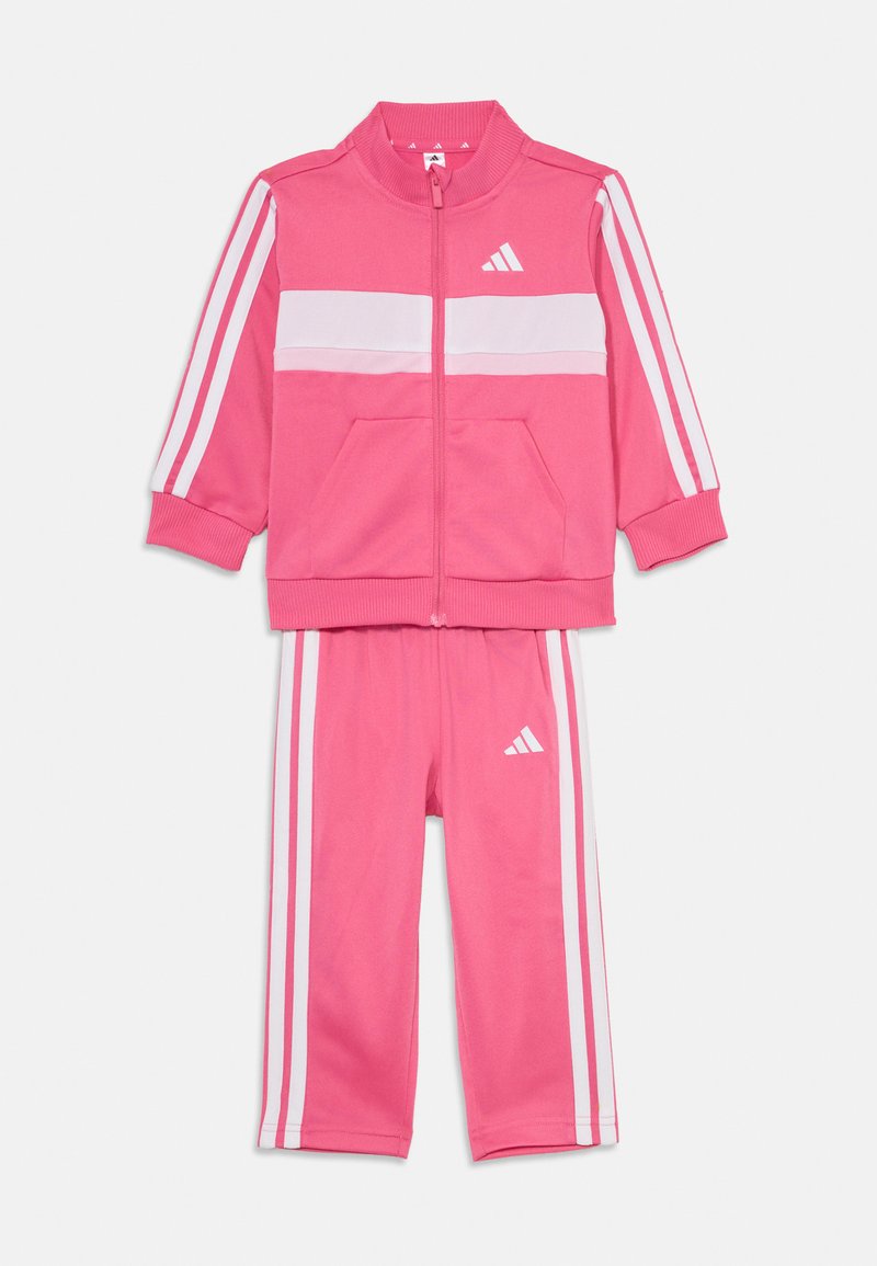 Rožinė Adidas treningo apranga, kurią sudaro zip-up striukė su baltais juostų akcentais ir atitinkamos kelnės, pagamintos iš lygios, elastinės medžiagos.