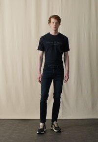 Black "Marc O'Polo" t-shirt, dark blue jeans, and black sneakers with beige accents. Model stands against a neutral backdrop.