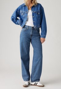 Denim jacket in faded blue with a classic collar, front pocket details, and long sleeves. Paired with high-waisted, loose-fitting blue jeans.