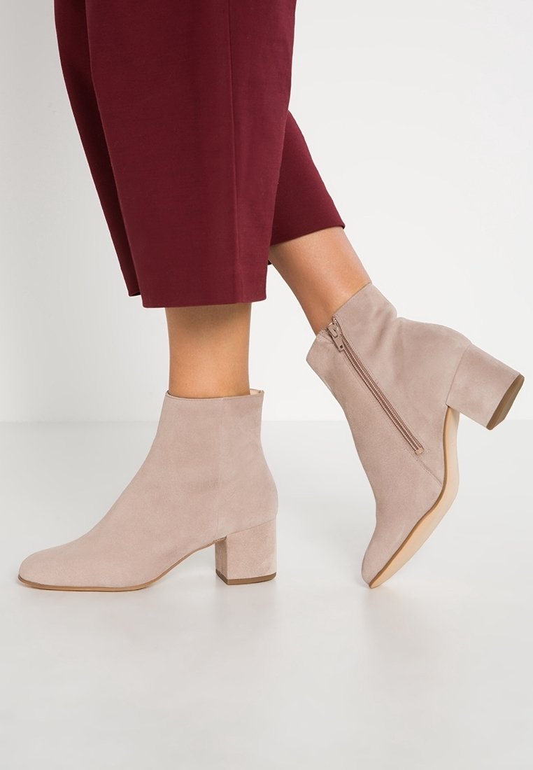 Femme portant des bottines en daim beige à talons blocs et fermetures éclair sur le côté, associées à un pantalon court bourgogne.