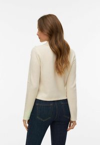 Krēmkrāsas adīts džemperis ar gariem piedurknēm un pērļu akcentiem, valkāts kopā ar tumši zilām pieguļošām džinsiem. Džempera dizains ir saīsināts ar apaļu kakla izgriezumu.