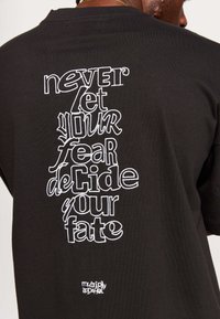 Black cotton t-shirt featuring white embroidered text on the back: "never let your fear deCide your fate" with a small logo at the bottom.