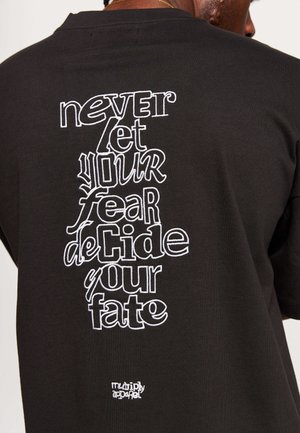 Camiseta de algodón negra con texto bordado blanco en la parte posterior: "nunca dejes que tu miedo decida tu destino" con un pequeño logotipo en la parte inferior.