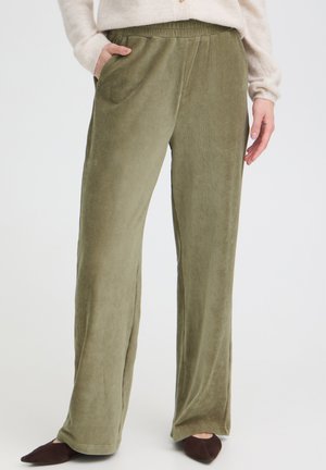 Femme portant un pantalon ample en velours côtelé vert olive avec une taille élastique et des poches latérales, associé à des chaussures marron à bouts pointus.