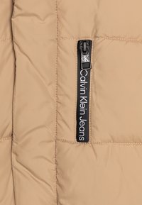 Beżowa kurtka puchowa o pikowanej fakturze. Posiada czarny zamek błyskawiczny i metkę z logo "Calvin Klein Jeans" po lewej stronie.