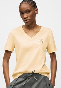 Γυναίκα που φοράει ανοιχτό κίτρινο T-shirt με V-λαιμόκοψη και λογότυπο "cK" στο στήθος και γκρι παντελόνι με κορδόνια, στέκεται μπροστά σε απλό λευκό φόντο.