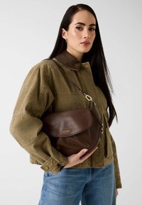 Bruine handtas van textuurleer met een klep en zilveren hardware, met het "GUESS"-logo, gedragen crossbody over een olijfgroene jas.