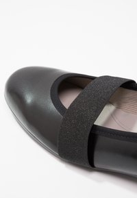 Chaussure plate Mary Jane noire avec bout arrondi et large bande élastique scintillante sur le cou-de-pied, sur fond clair.