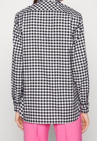 Chemise à carreaux noir et blanc avec un col classique, des manches longues et des poignets boutonnés. Le tissu présente un motif texturé. L'ourlet est légèrement arrondi.