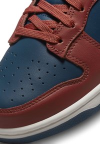 Sneaker in pelle con tomaia blu e dettagli perforati, sovrapposizioni rosse e lacci marroni testurizzati. Suola intermedia bianca e suola esterna blu.