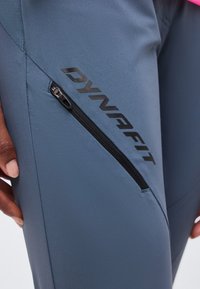 Κοντινό πλάνο σε μπλε αθλητικό παντελόνι με μαύρο λοξό φερμουάρ και λογότυπο "DYNAFIT", που φοριέται από άτομο με το χέρι κοντά στον μηρό.